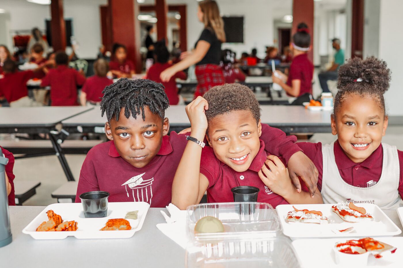 3 kids sit together smiling and enjoying lunch in the cafeteria
